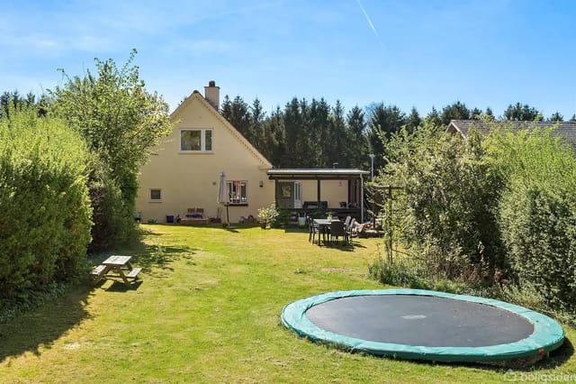 A yellow-painted house stands on a lawn with a trampoline in the foreground. There are garden furniture next to the house, surrounded by bushes and trees under a blue sky.