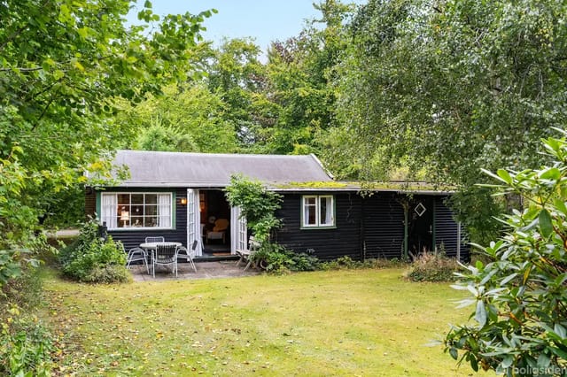 A black wooden cabin surrounded by lush, green vegetation. In front, a table with chairs sits on a small terrace, all framed by a lawn and dense forest.