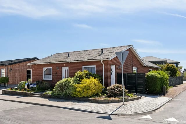 Single-story red brick house with flat roof on a corner lot, surrounded by green bushes and flower beds in sunshine.