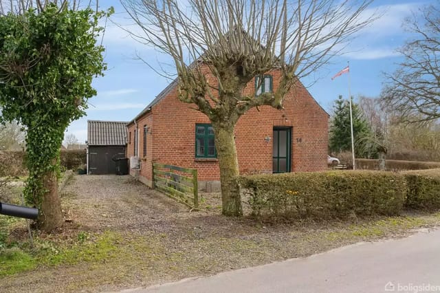 A red brick house stands quietly by a gravel road. A large, trimmed tree foreground surrounded by bushes. Danish flag waving in the background. Dark outbuilding seen behind the house.