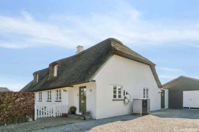 White thatched house with wooden fence and ceramic pots at the entrance; located on a gravel plot in sunny weather. House number 10 is shown by the door.
