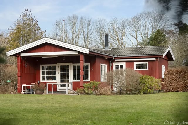 Red wooden house with terrace in a garden surrounded by bushes and trees. Chimney pipe on the roof. Lawn in front of the house.