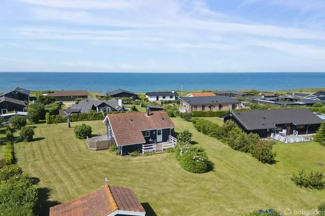 A summer house with a reddish-brown roof surrounded by a well-kept lawn and scattered bushes. It is located in a residential area with a view of the sea under a blue sky.