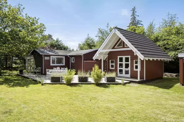 A red wooden house with white details stands on a green lawn surrounded by trees. In front of the house is a terrace with plants and garden furniture.