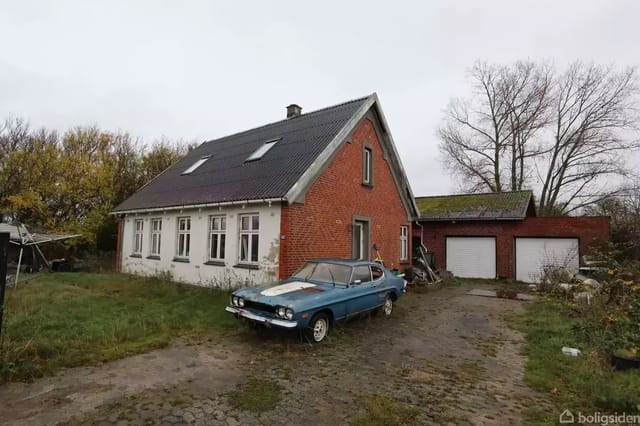 A vintage red brick house with a sloped roof stands by a weed-covered driveway. A blue vintage car is parked in front of garage doors in the background. The atmosphere is abandoned and cluttered.