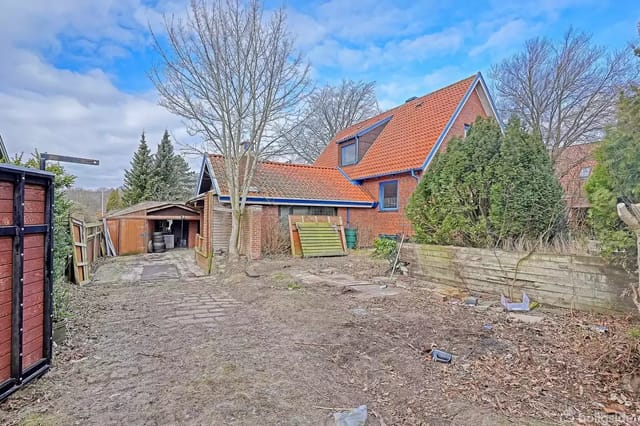 House with red tile roof and brick walls, surrounded by trees and bushes in a garden. A shed appears to the left. The sky is slightly cloudy.