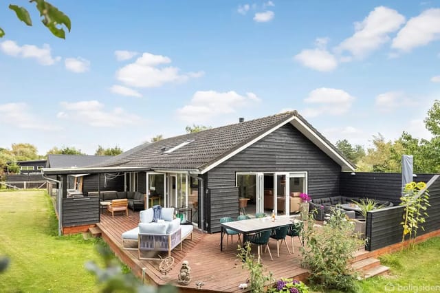 Black wooden house with large windows on a wooden terrace, furnished with white and blue seating, surrounded by green plants and grass. Blue sky with a few clouds in the background.