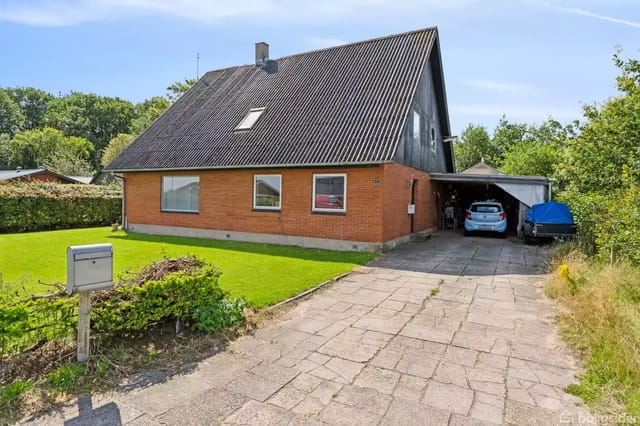 A red brick house with a black roof sits by a paved driveway. A carport to the right houses two cars. Green bushes and trees surround the house.