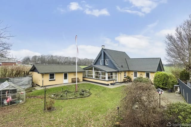 A yellow house with a black roof stands in a green garden. A flagpole is by the house, surrounded by bushes and trees. A small glass structure is seen to the left.