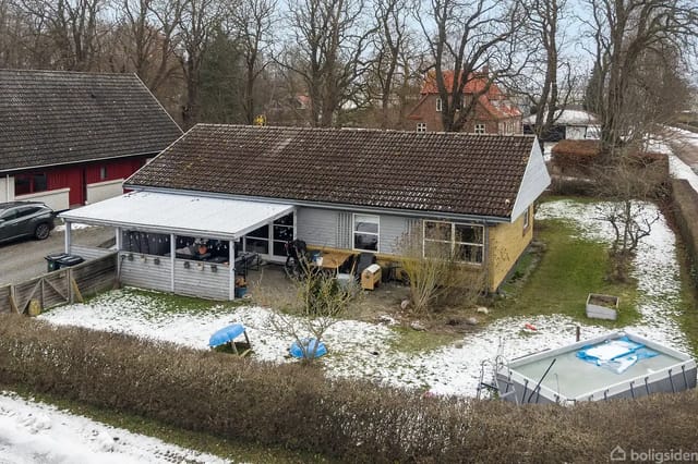 A villa stands in a garden with light snow cover. There is a carport next to the house and a swimming pool in the garden. Trees surround the area.