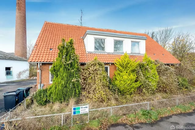 The house has a red tile roof and is surrounded by tall green bushes. There are waste containers next to it, and a sales poster is visible in the yard.