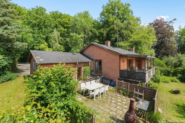 Brick house with terrace, surrounded by lush garden and trees. In the foreground, a terrace with dining table, chairs, outdoor fireplace, and grill.