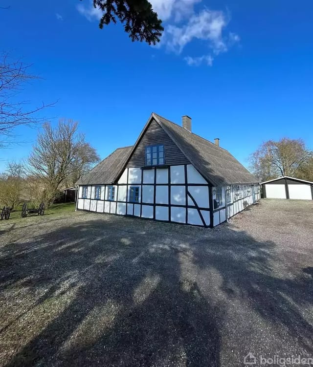 Timber-framed house with thatched roof on a gravel courtyard, surrounded by trees and a clear blue sky in a rural, peaceful setting.