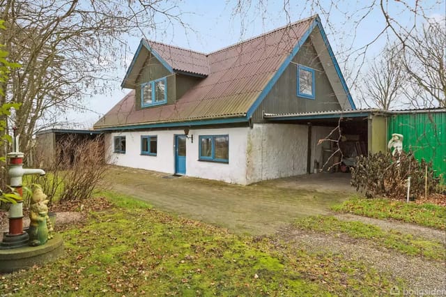 A two-story house with blue windows and door on a plot with lawn and trees. Next to the house is a carport and a decorative water pump with garden gnomes.