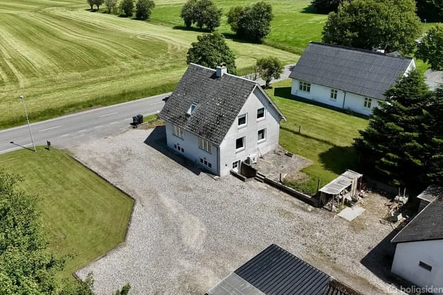 A white house with a gray roof ridge stands on a gravel driveway. The background shows green fields and another white building.