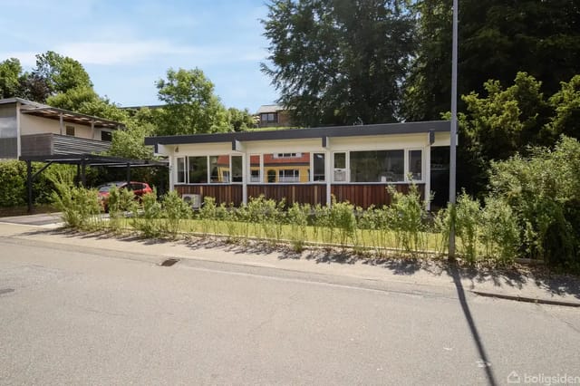A low, modern building with large windows surrounded by green bushes, located in a residential area with trees and other houses in the background.