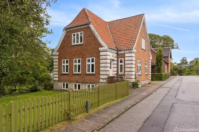 Red brick house with a pointed red tile roof, located along a road. The house has white window frames and is surrounded by a green-painted wooden fence and a garden full of trees.