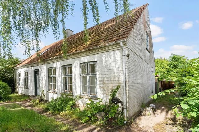 An old white brick house with a red tile roof, partially covered with moss, stands quietly in a lush garden with green bushes and hanging branches under a blue sky.