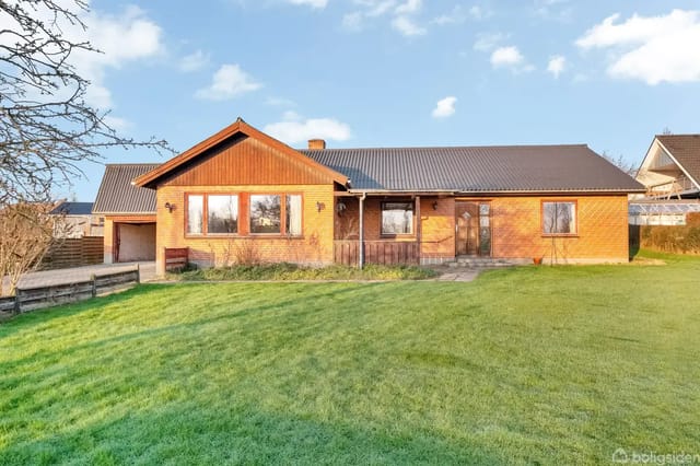 A brown brick villa with a gray roof stands on a well-kept lawn. Large windows and a main door are visible. Trees surround the house under a blue sky with a few clouds.
