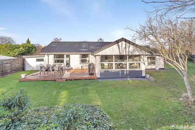 A house with a flat roof stands quietly in a lush garden surrounded by trees. A wooden terrace with garden furniture is in front of the house.