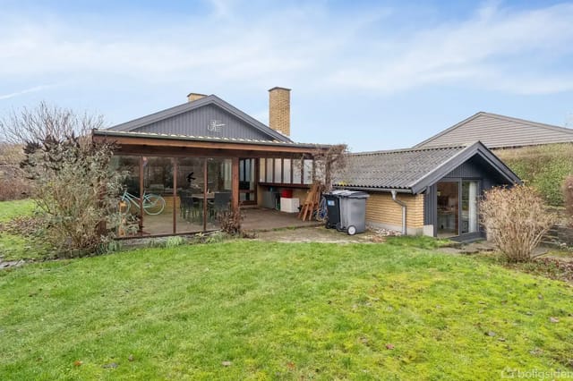 A modern house with large glass sections facing a well-maintained lawn. Bicycles are stored on the terrace, surrounded by bushes and neighboring roofs in the background.