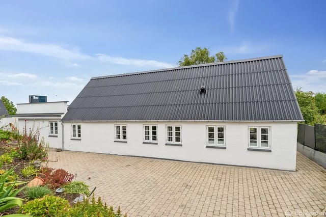 Traditional white house with sloped black roof, surrounded by cobblestone courtyard and landscaped flower beds under a clear blue sky.