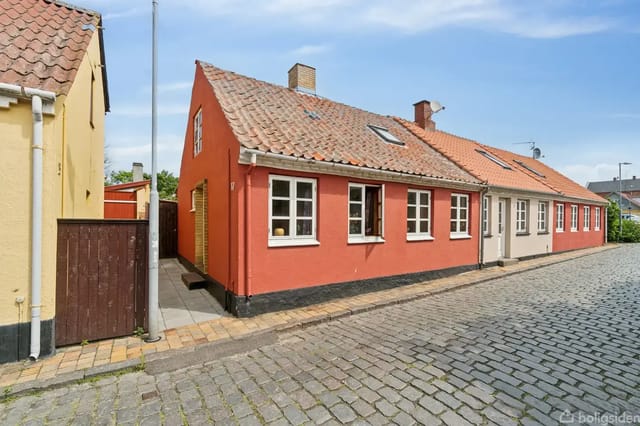 Red brick house with sloped tiled roof along a cobblestone street. White windows and a brown wooden gate. Clear, sunny day.