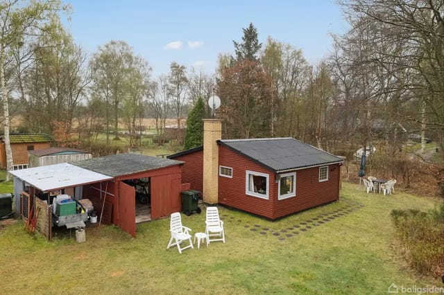 A small, red wooden house with a chimney stands in a grass-covered garden. Next to it is a shed with open doors. There is garden furniture around and surrounding trees.