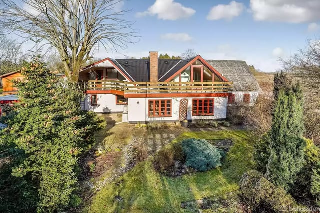 A house with a red ridge and a balcony extends over two floors, surrounded by a lush garden with trees and bushes under a blue sky.