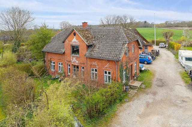 An older red brick house stands empty, surrounded by green bushes and trees. Several cars are parked on a gravel driveway next to the house. The surrounding landscape is rural.