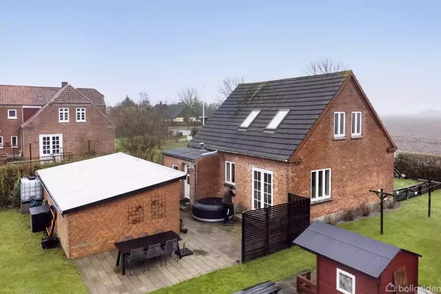 A red brick house with a sloped roof and several windows surrounded by a green garden. In front of the house is a terrace with a table and chairs. Another building is nearby.