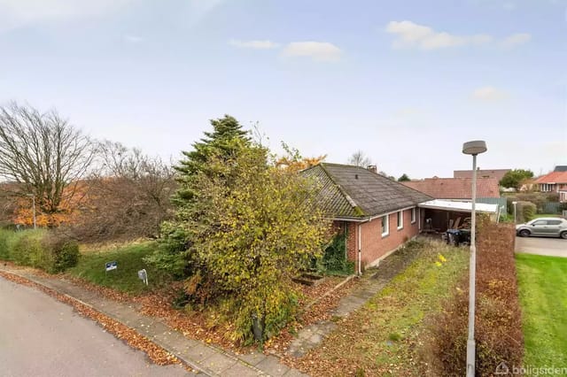 A red brick house in a residential neighborhood surrounded by trees and a well-kept lawn. A driveway in front, with other houses and a parked car in the background.
