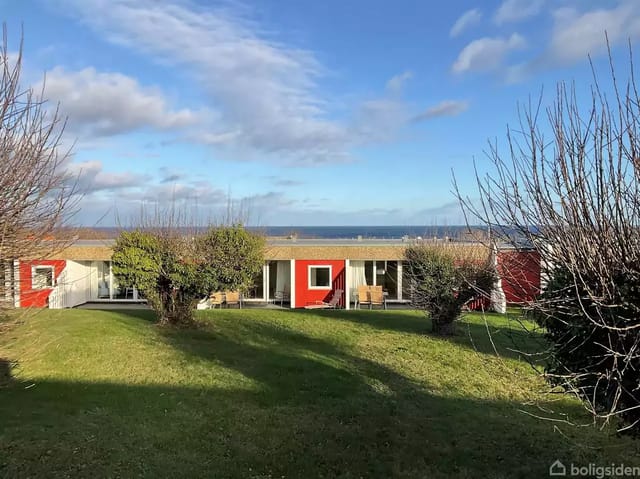 A modern building with red-white facades stands in a green garden with trimmed bushes, while the sea is visible in the background under a cloudy, blue sky.