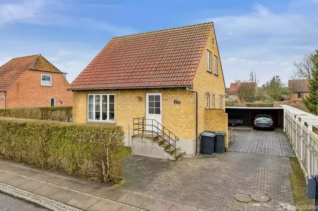 Yellow brick house with red tile roof next to a driveway and carport, surrounded by a hedge.