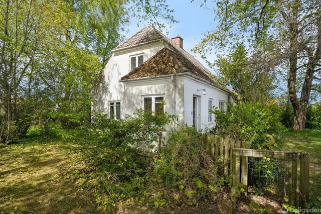 A white house with a steep brown roof sits quietly in a lush garden surrounded by green trees and bushes. A low wooden gate marks the entrance to the garden.