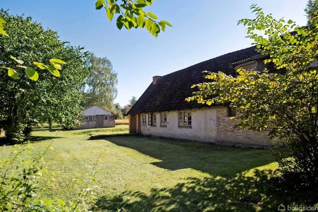 A traditional half-timbered house stands quietly in a sunny garden surrounded by green trees. In the background, a smaller building is visible on the large, open lawn.
