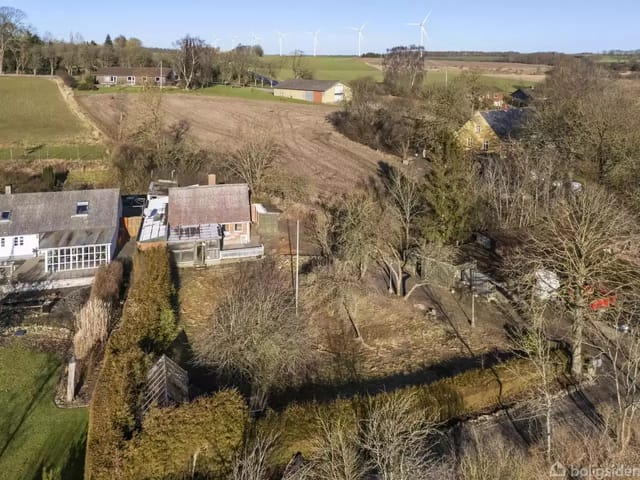 House facade facing a grassy garden with trees; the surroundings are rural fields and wind turbines in the distance under a clear sky.