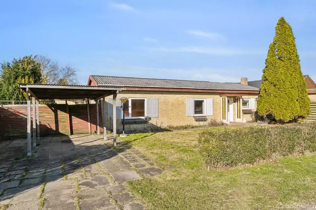 A standalone, low, brick house with several small windows and a white door. In front of the house stands a tall, slender bush, and there is a paved driveway and a small carport beside it.