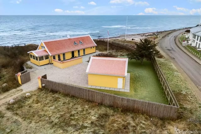 A yellow house with a red tile roof stands quietly by a coast. In the background, there is sea, a road, and other buildings. A small gravel yard and lawn surround the house.