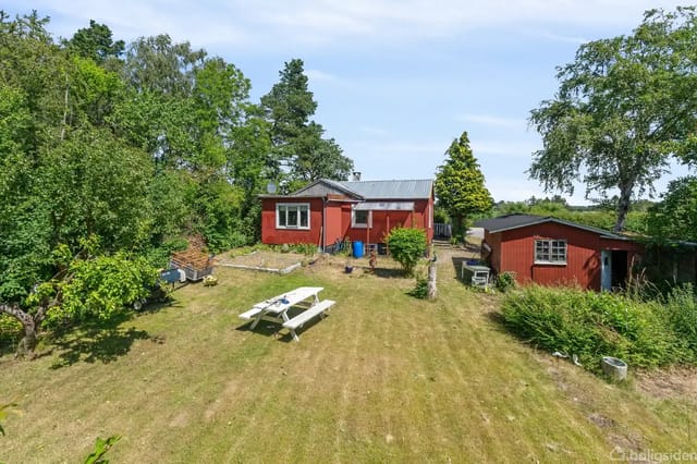 A red house stands quietly in a green garden with a white picnic table in the foreground, surrounded by tall trees and bushes under a blue sky.