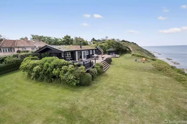 A black summer house with a terrace surrounded by lush bushes stands on a large lawn by the coast. In the background, the sea and a rocky shoreline are visible.