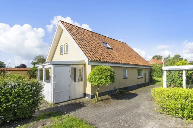 A yellow house with a red tiled roof stands in the sunshine. The house has a small conservatory and a surrounding garden, with a paved path leading to the entrance door.