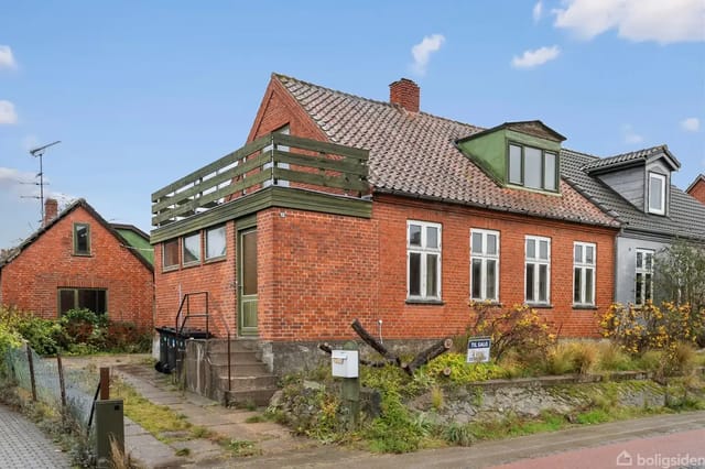 Red brick house with roof terrace, located on a residential street surrounded by other houses and sparse bushes. The image clearly shows the house's facade details and garden.