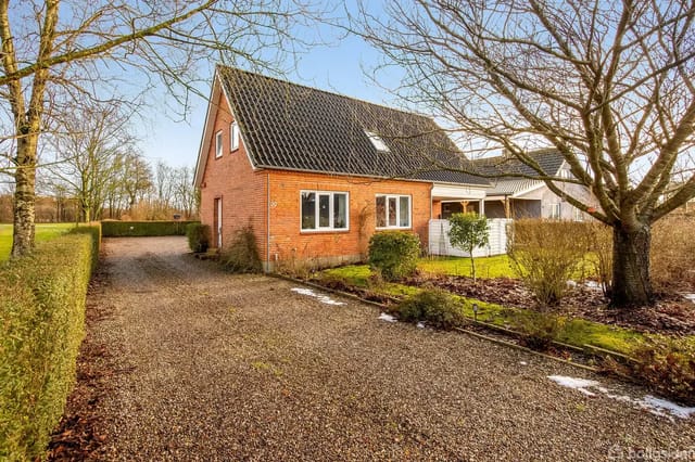 Red brick house with black roof by a gravel road, surrounded by a well-kept garden and some trees. A clear blue sky in the background.