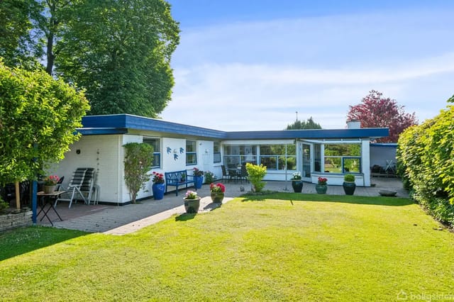 A white bungalow with blue roof trim stands in a well-kept garden with a green lawn. Potted plants decorate the courtyard. Trimmed bushes surround the area.