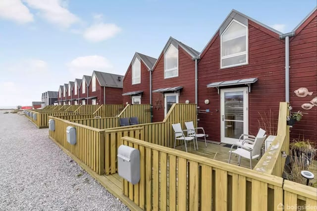 Red houses with sloping roofs lined up along a gravel road. Each house has a small terrace with chairs, and there are trash bins by the railing. Blue sky in the background.