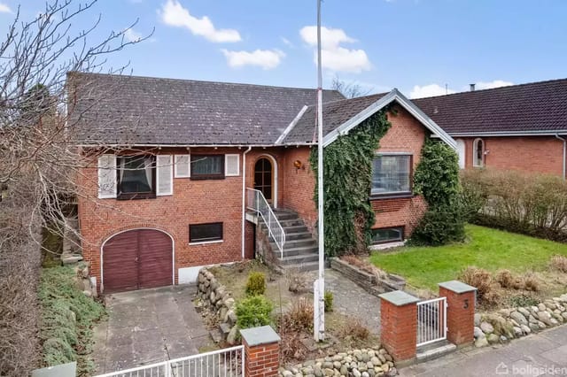 A brick villa with a garage door and green plants growing up the facade. The entrance has a staircase, and the house is surrounded by a garden with a lawn and bushes in a residential area.