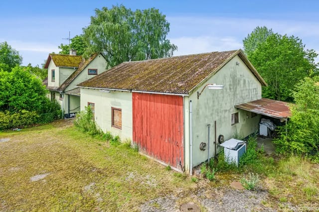 An old barn with a red door stands in a courtyard surrounded by trees and grass. The building appears worn with moss on the roof.