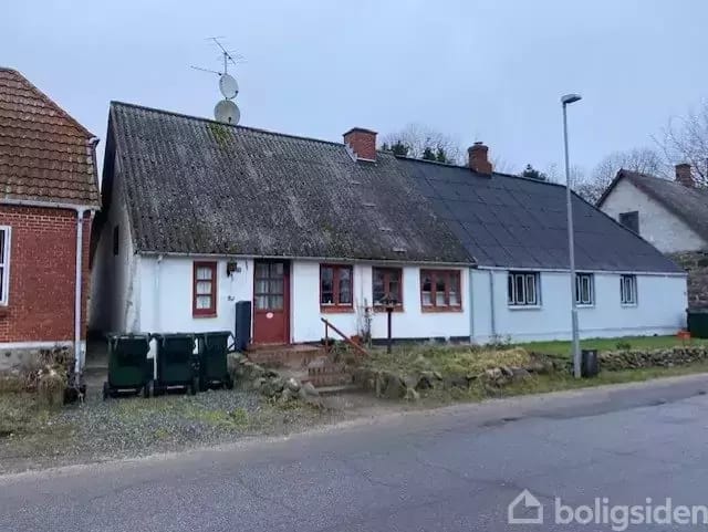 An old white terraced house with a black roof in a village. There is a red front door with three small steps in front, surrounded by other traditional buildings.