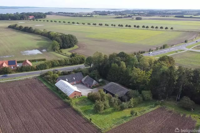 A farm with several connected buildings surrounded by fields and forest. In the background, a country road and open landscape are visible.